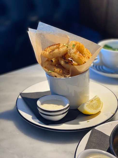 a photo of a serving of Calamari from Pizza Express. It is on a plate with a side dish of dressing and a wedge of lemon